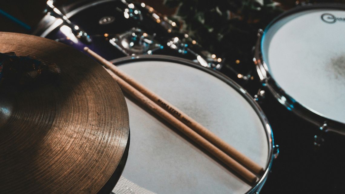 Close-up of drumsticks resting across a snare drum beside a cymbal, captured in a dark practice studio to ground music culture, sustainable merch storytelling, and organic cotton streetwear relevance. Above Average Clothing