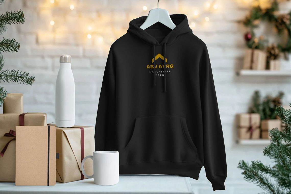 Black organic cotton hoodie on a hanger with gold ascent caret logo, shown in a UK studio setting with gifts and a reusable bottle, highlighting sustainable merch and circular, purpose-led streetwear from Manchester.